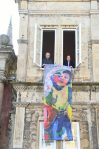 Zwei Personen präsentieren eine farbenfrohe Fahne vor einem Fenster.