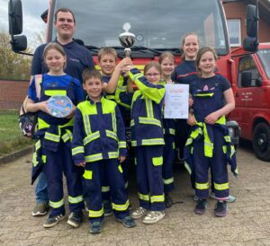 Eine Gruppe von Kindern in Feuerwehrkleidung hält einen Wanderpokal und eine Urkunde hoch.