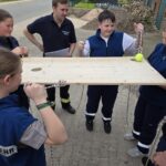 Kinder halten mit Seilen ein Holzbrett, auf dem ein Tennisball liegt.