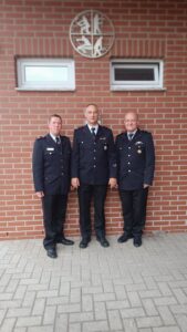 Drei Männer in dunkelblauen Feuerwehruniformen stehen vor einer roten Backsteinwand.