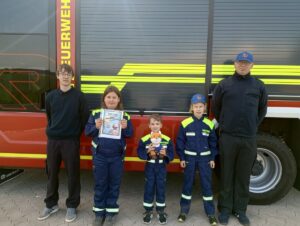 Kinder in Feuerwehrkleidung vor einem Löschfahrzeug, ein Kind hält einen Stofffuchs im Arm.