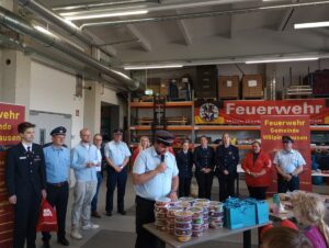 Eine Gruppe von Personen bei einer Siegerehrung in einer Feuerwache; ein Mann spricht in ein Mikrofon, im Vordergrund stehen Preise.