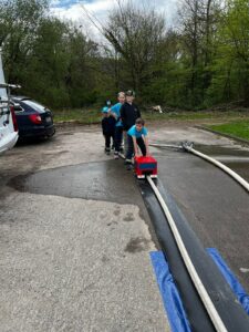 Kinder in Feuerwehrkleidung an einer Station mit einem Feuerwehrschlauch und einer roten Kiste.