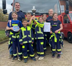 Gruppe von Kindern in Feuerwehrkleidung mit Wanderpokal und Urkunde vor einem Einsatzfahrzeug.