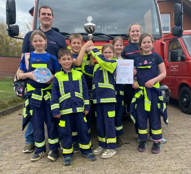 Gruppe von Kindern in Feuerwehrkleidung mit Wanderpokal und Urkunde vor einem Einsatzfahrzeug.