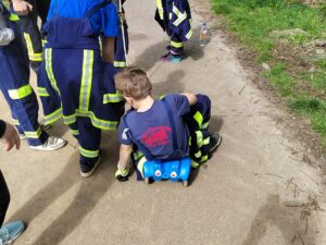 Ein Kind in Feuerwehrkleidung sitzt auf einem blauen Bobbycar auf einem Waldweg.