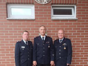Drei Männer in dunkelblauen Feuerwehruniformen stehen vor einer roten Backsteinwand.
