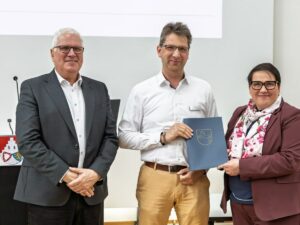 Drei Personen stehen vor einer Projektionsfläche, eine Person hält einen blauen Ordner mit einem Wappen.